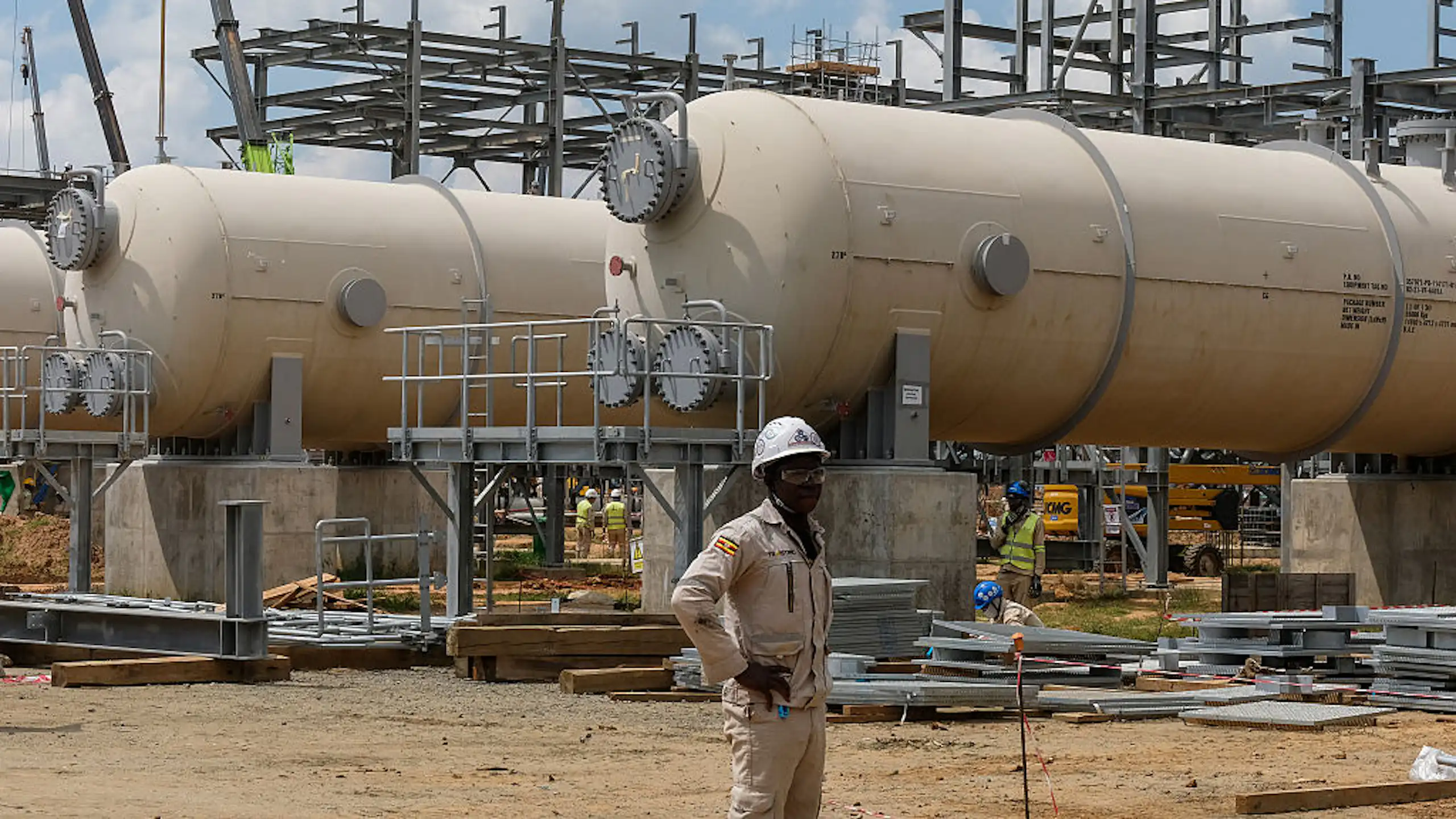 Construction workers stands next to large gas tanks.