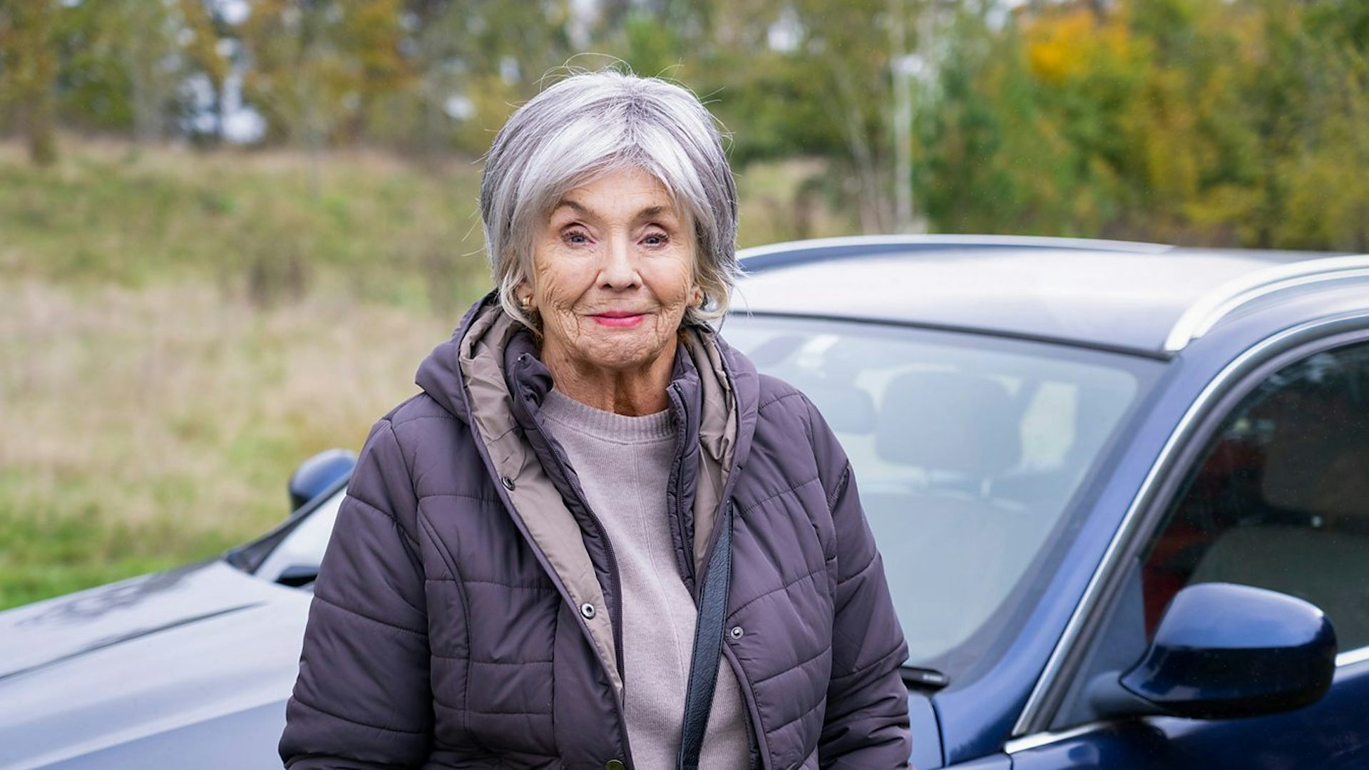 Sue Johnston as Sue standing in front of a car.