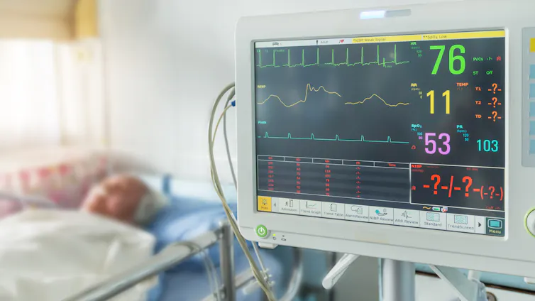 Sala de un hospital con un monitor donde aparecen cifras u gráficos de las constantes vitales y un paciente al fondo
