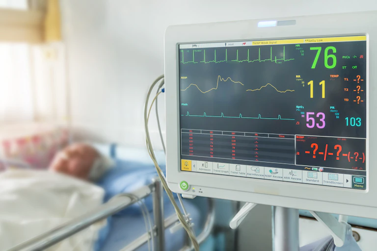 Sala de un hospital con un monitor donde aparecen cifras u gráficos de las constantes vitales y un paciente al fondo