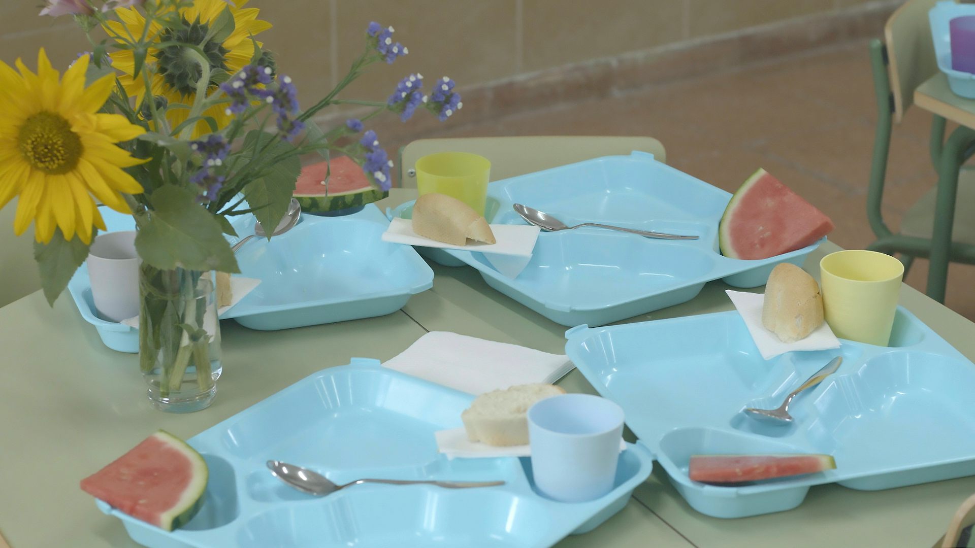 Mesa de comedor escolar preparada con bandejas azules, vasos y porciones de sandía antes del servicio.