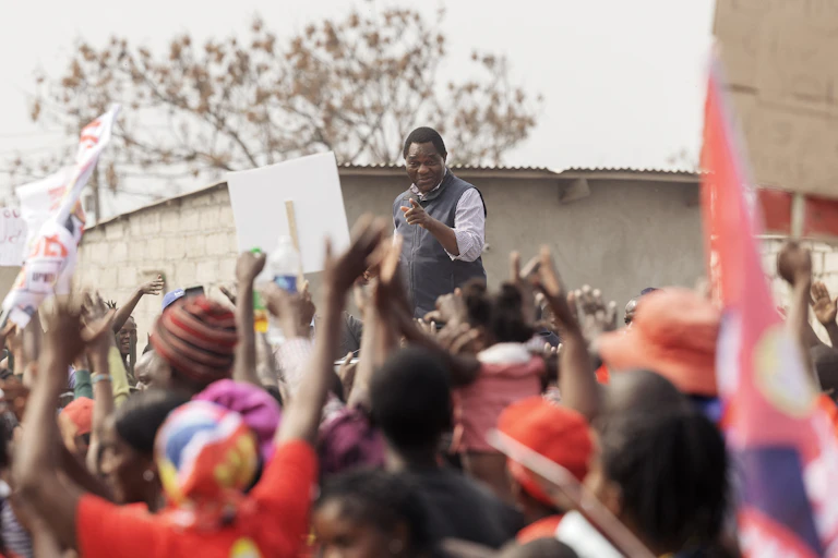 An African man in a smart shirt and waistcoat stands above a crowd of people who wave their arms and hold placards.