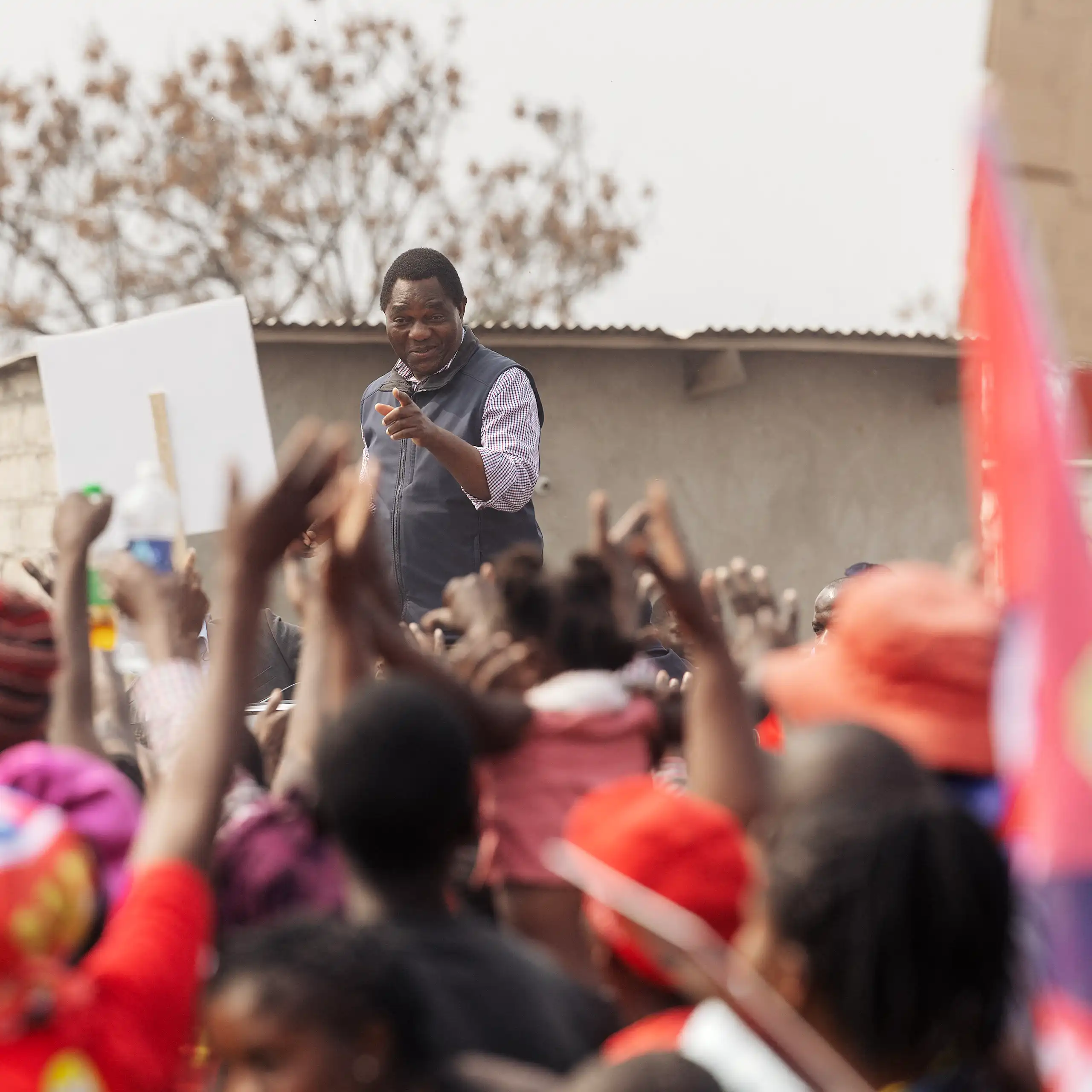 An African man in a smart shirt and waistcoat stands above a crowd of people who wave their arms and hold placards.