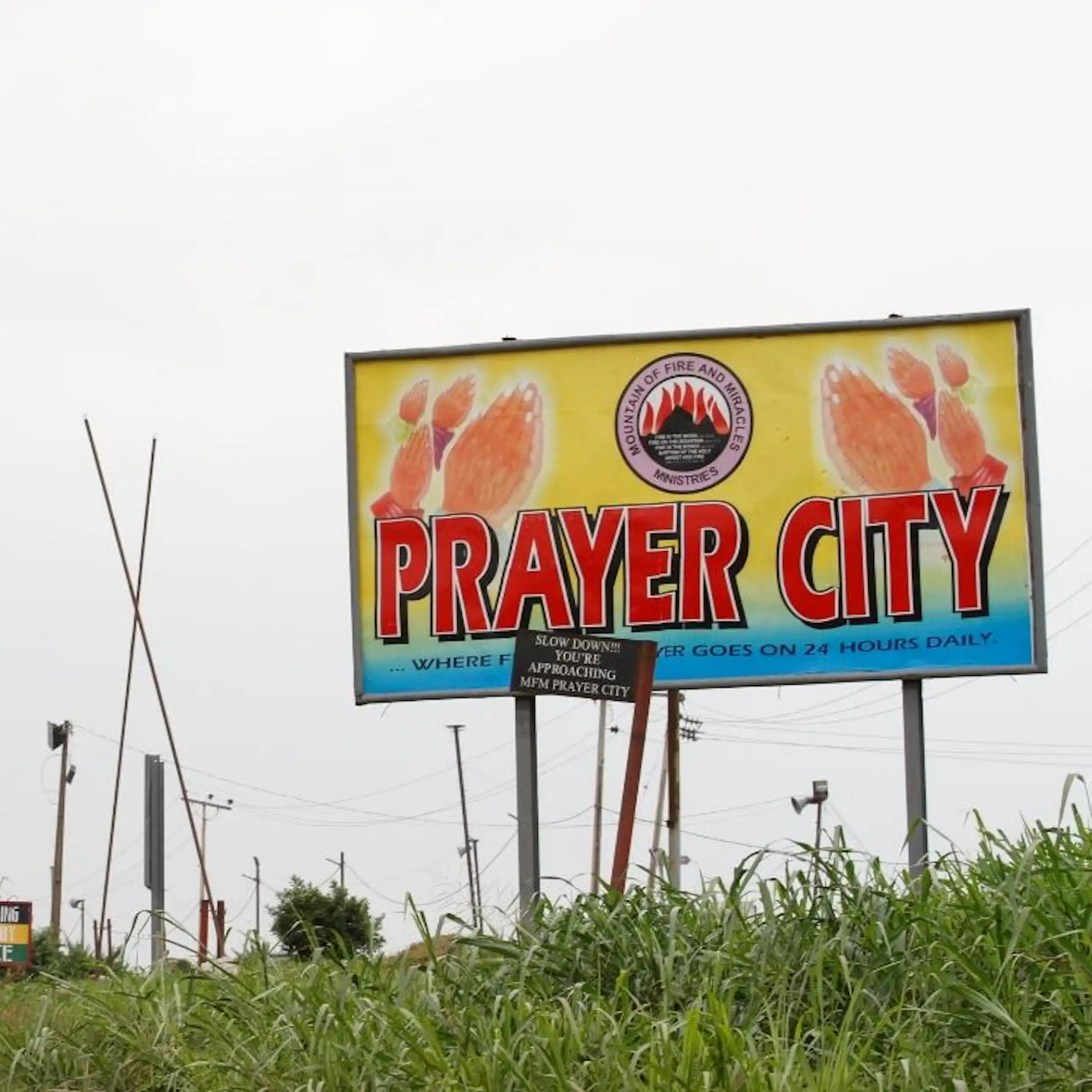 Cars drive past a large bright yellow and red billboard that reads "Prayer City".