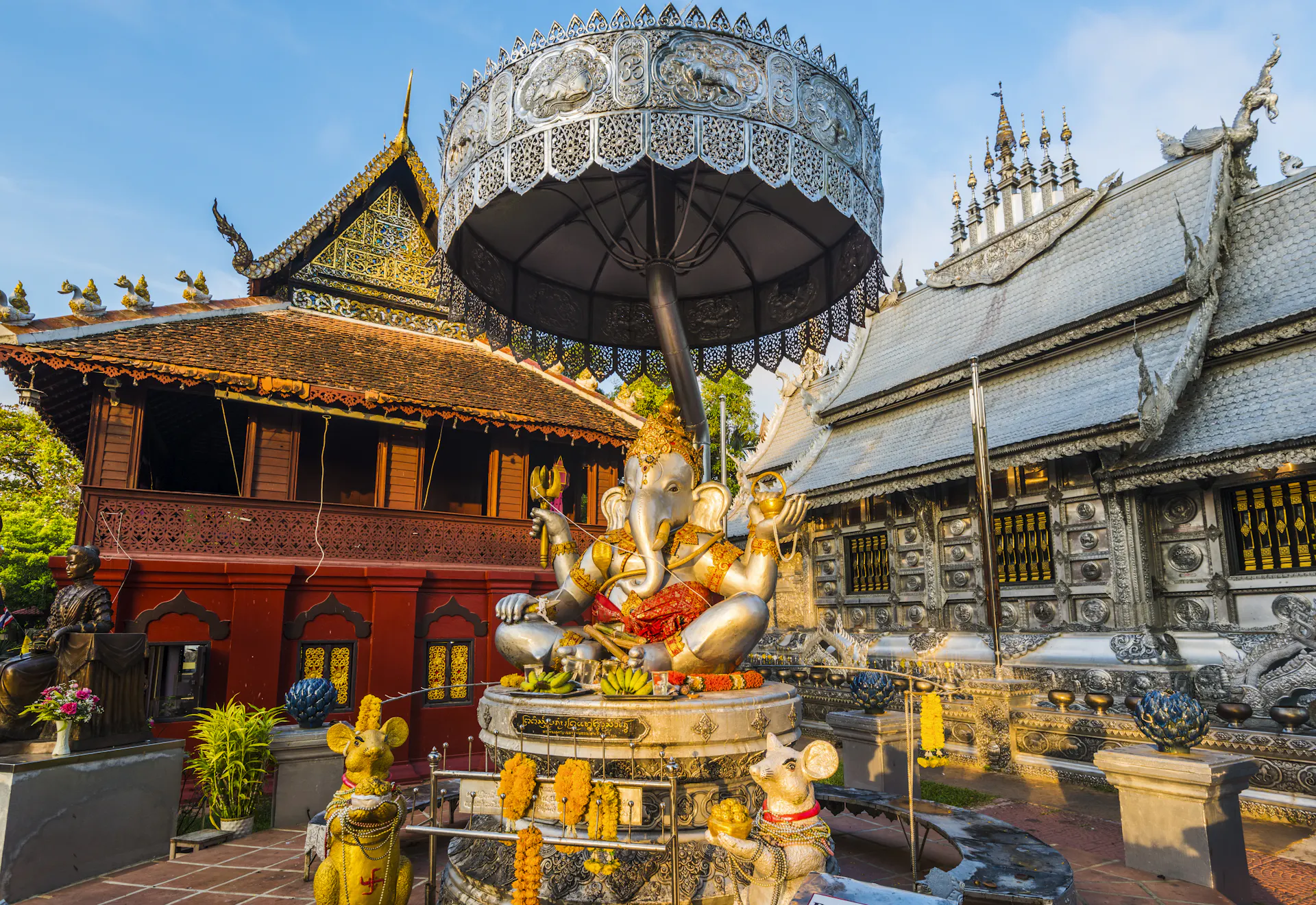 A large statue of the elephant-headed god Ganesha with a silver umbrella above it and two mouse statues nearby, in the courtyard of a temple with colorful red walls.
