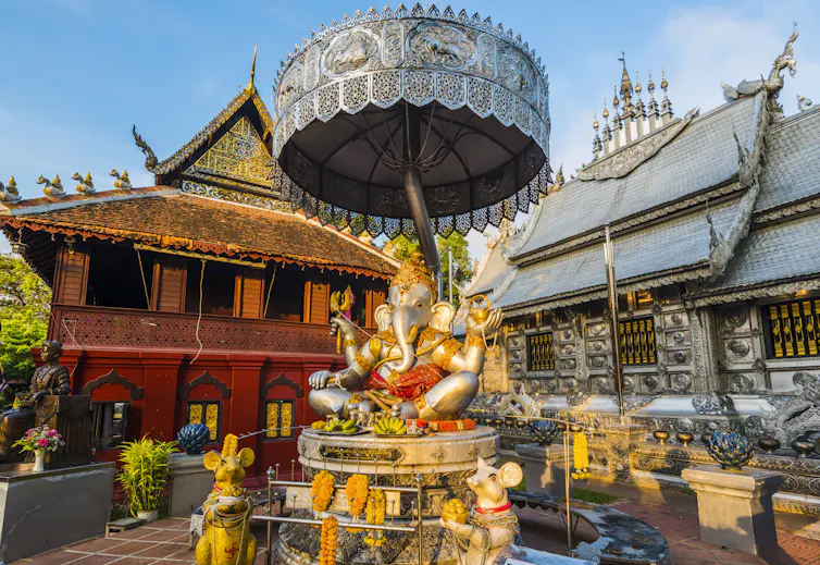 A large statue of the elephant-headed god Ganesha with a silver umbrella above it and two mouse statues nearby, in the courtyard of a temple with colorful red walls.