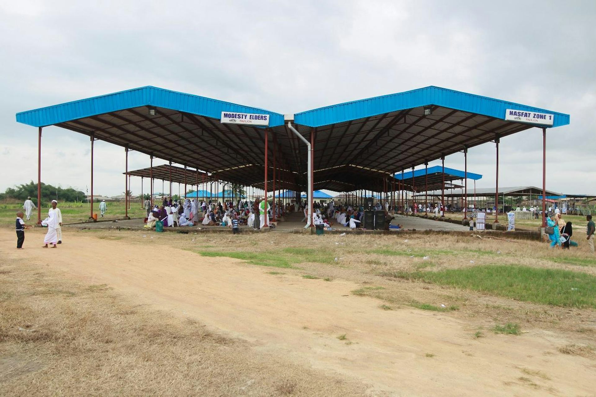 Many large blue roofs with people dressed in white congregating beneath them outdoors.