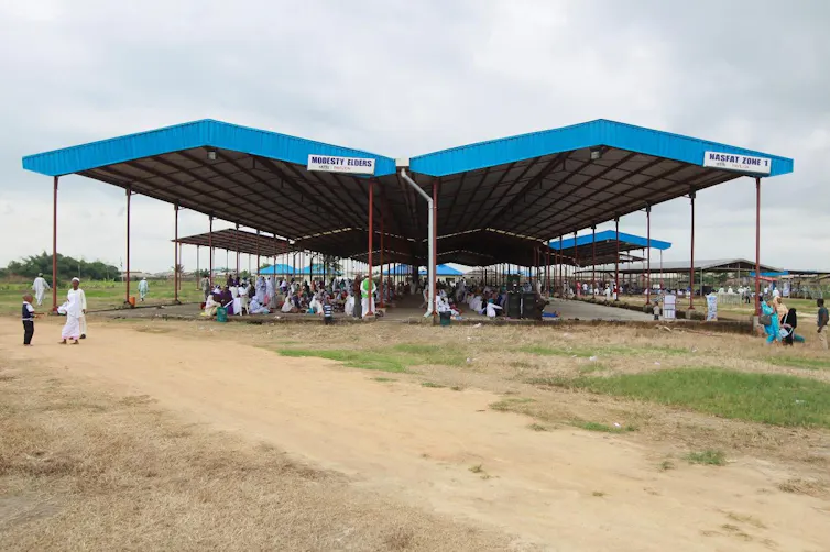 Many large blue roofs with people dressed in white congregating beneath them outdoors.