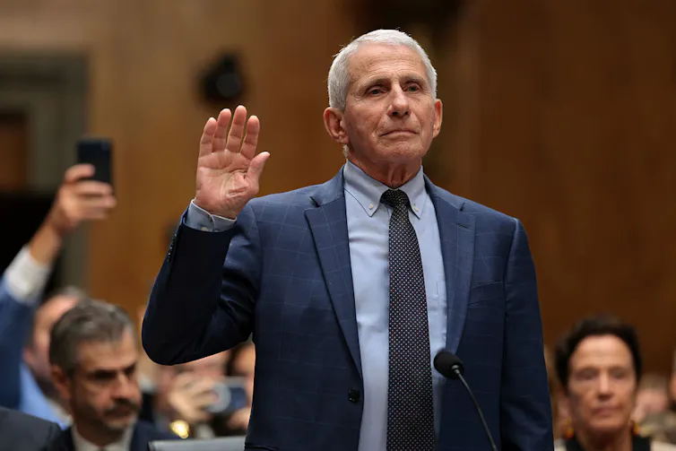 A man with white hair and wearing a suit raises his right hand in a courtroom.