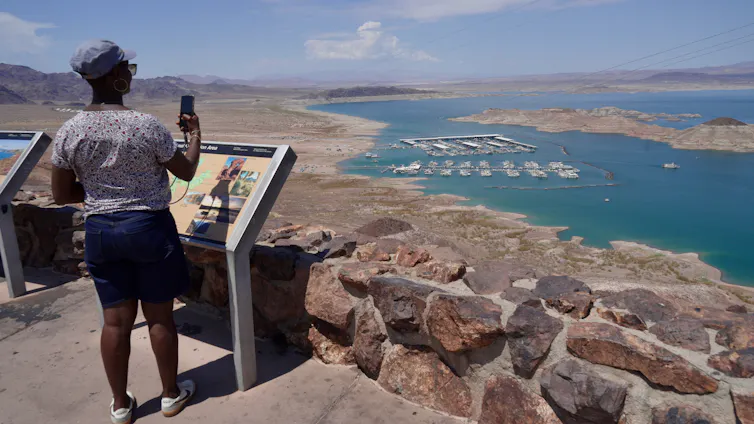 A person stands at an overlook, from which a lake is visible in the distance, with a wide dry area all around it.