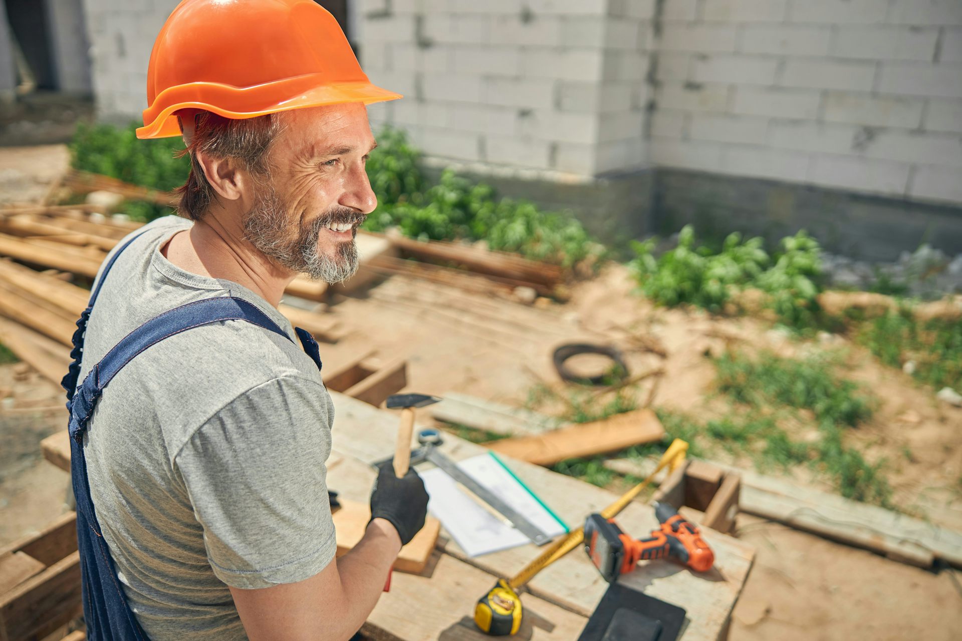 A man with a grey beard wearing overalls and an orange construction helmet on a construction site