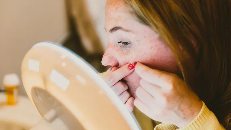 Young woman sitting at make-up mirror, squeezing pimple