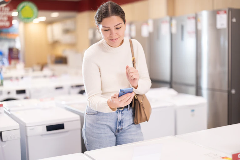 woman in white jumper blue jeans holds phone and scans label on washing machine in showroom