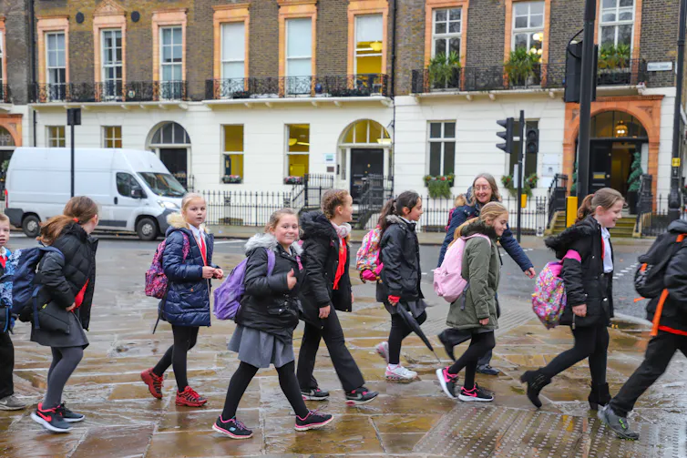 A group of schoolchildren wearing uniforms and winter coats walk along a road in Westminster with their teachers.