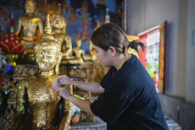 A woman in a blue shirt places a small sheet of gold leaf on a golden Buddha statue.
