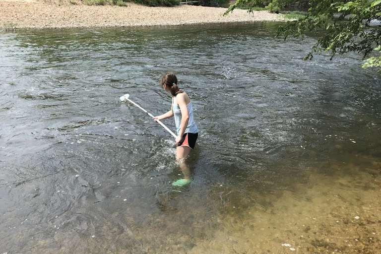 A person stands part of the way into a stream while holding a pole with a plastic bottle at the end.