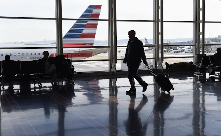 A shadowed traveler pulling a suitcase through an airport, with the tail of an airplane visible out the window.