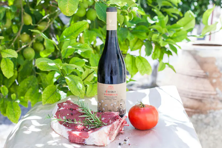 A bottle of wine on a table with a raw steak and a tomato.