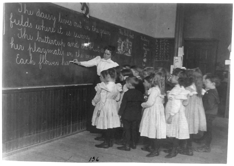 A black and white photo shows a group of children standing near a woman in a white shirt at a large blackboard that is covered in white chalk cursive writing.