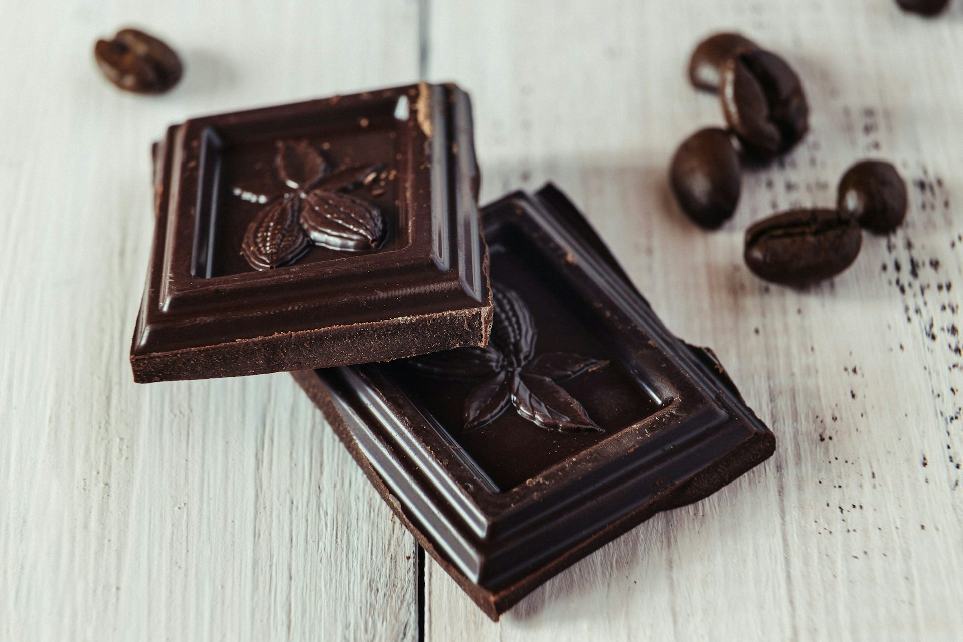 Two cubes of dark chocolate balanced one on the other, with a scattering of cocoa beans.