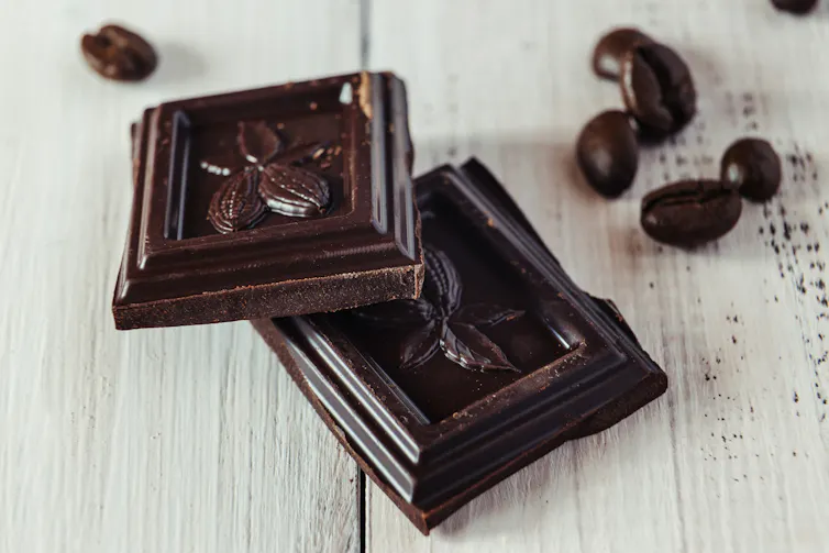 Two cubes of dark chocolate balanced one on the other, with a scattering of cocoa beans.