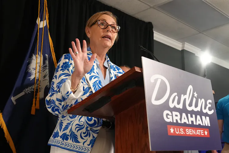 Sen. Darline Graham speaks from behind a lectern bearing a 'Darline Graham U.S. Senate' campaign sign.