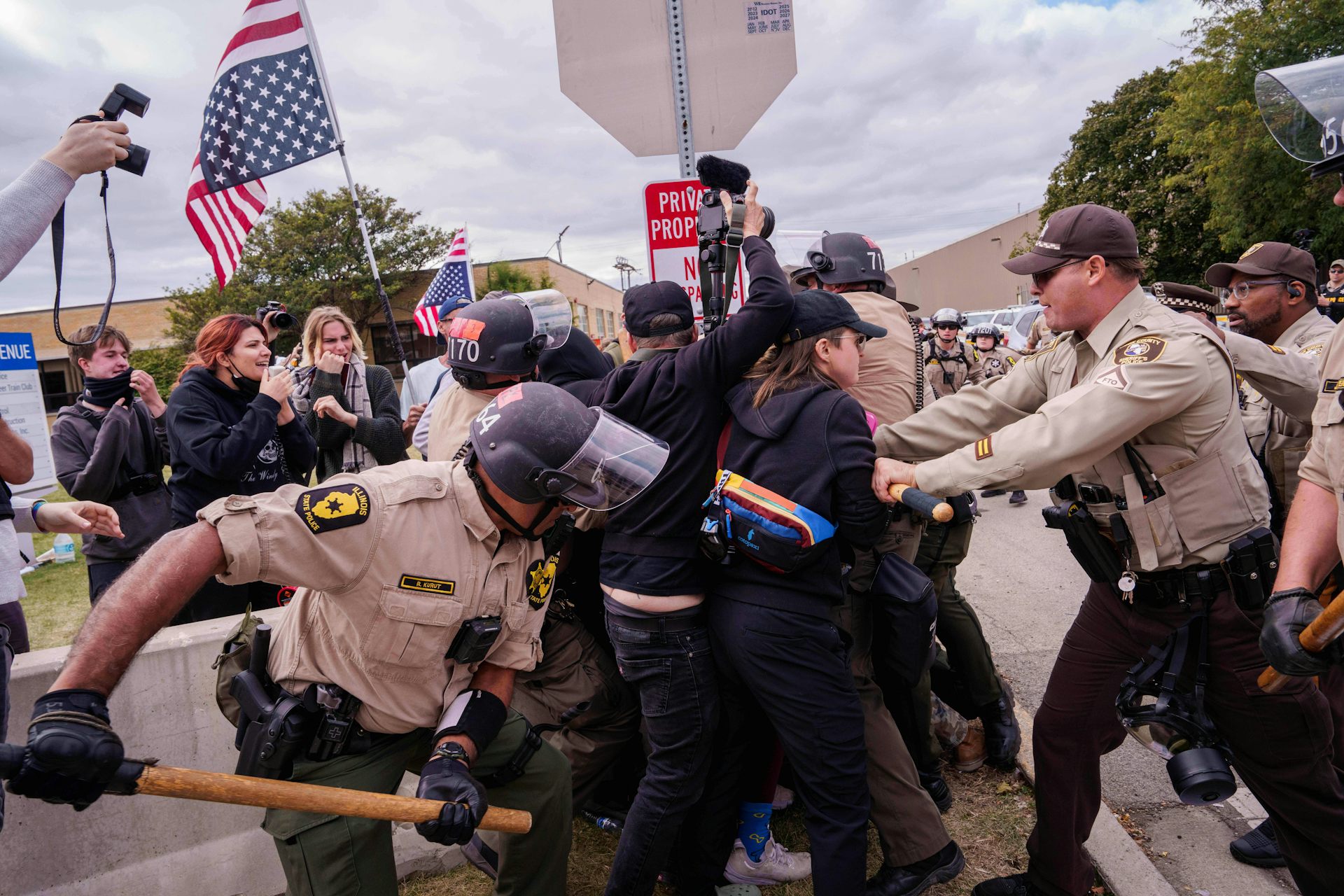 Uniformed officers in helmets grapple with protesters as onlookers record the scene with cameras.
