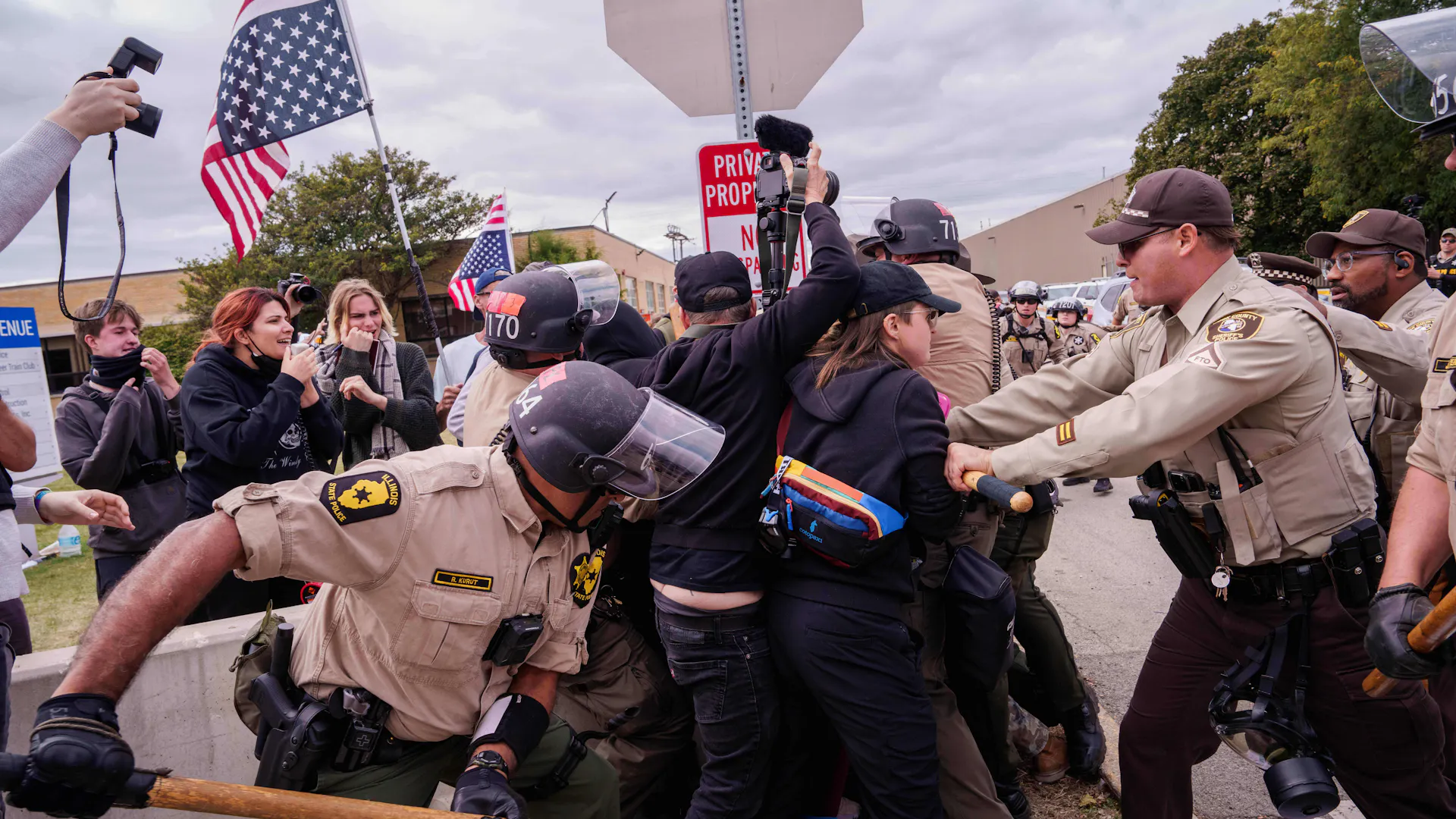 Uniformed officers in helmets grapple with protesters as onlookers record the scene with cameras.