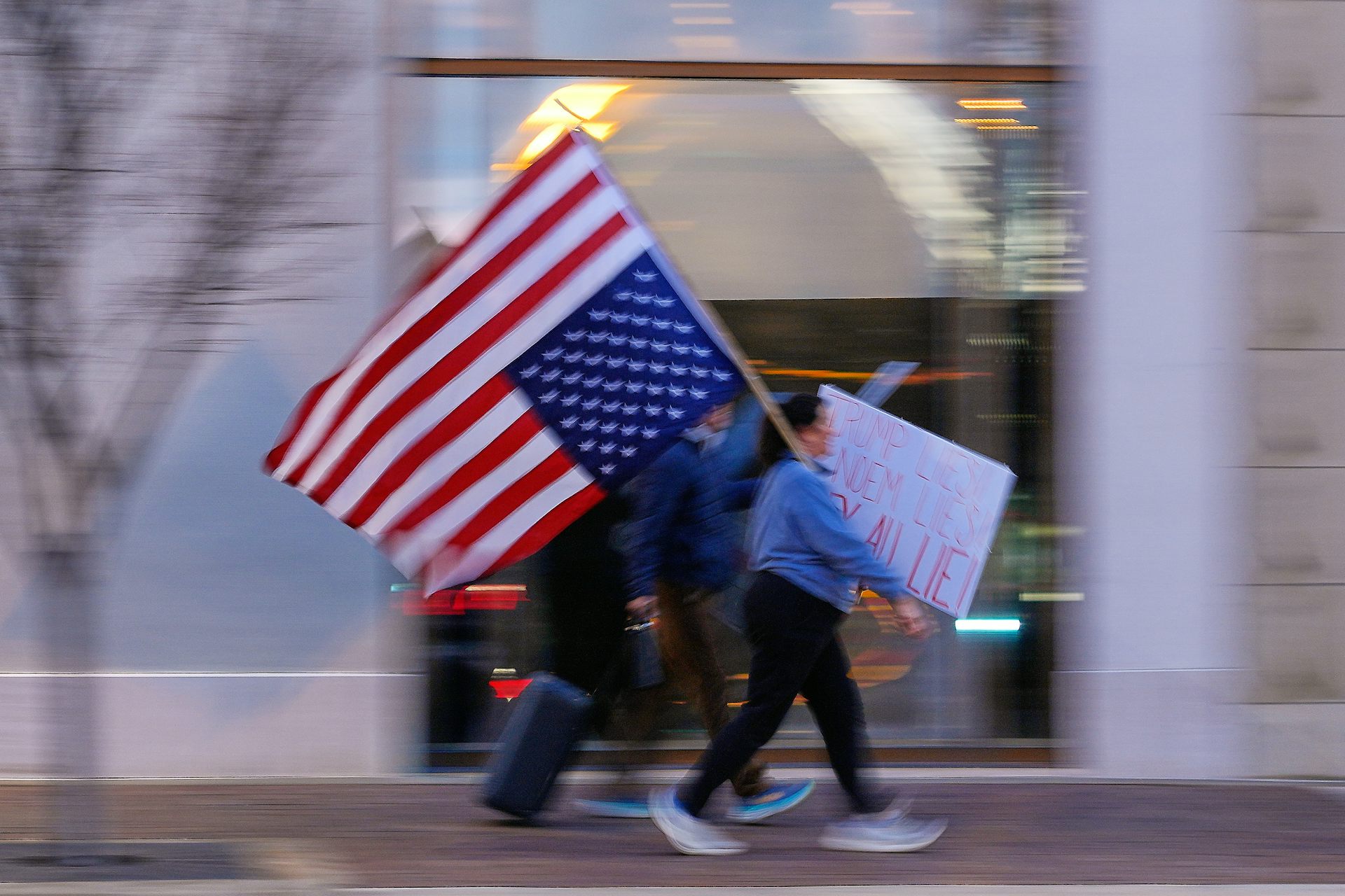 Blurry shot of protesters walking, one carrying a large upside-down American flag and another holding a cardboard sign