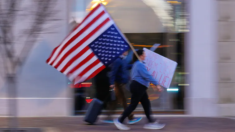 Blurry shot of protesters walking, one carrying a large upside-down American flag and another holding a cardboard sign