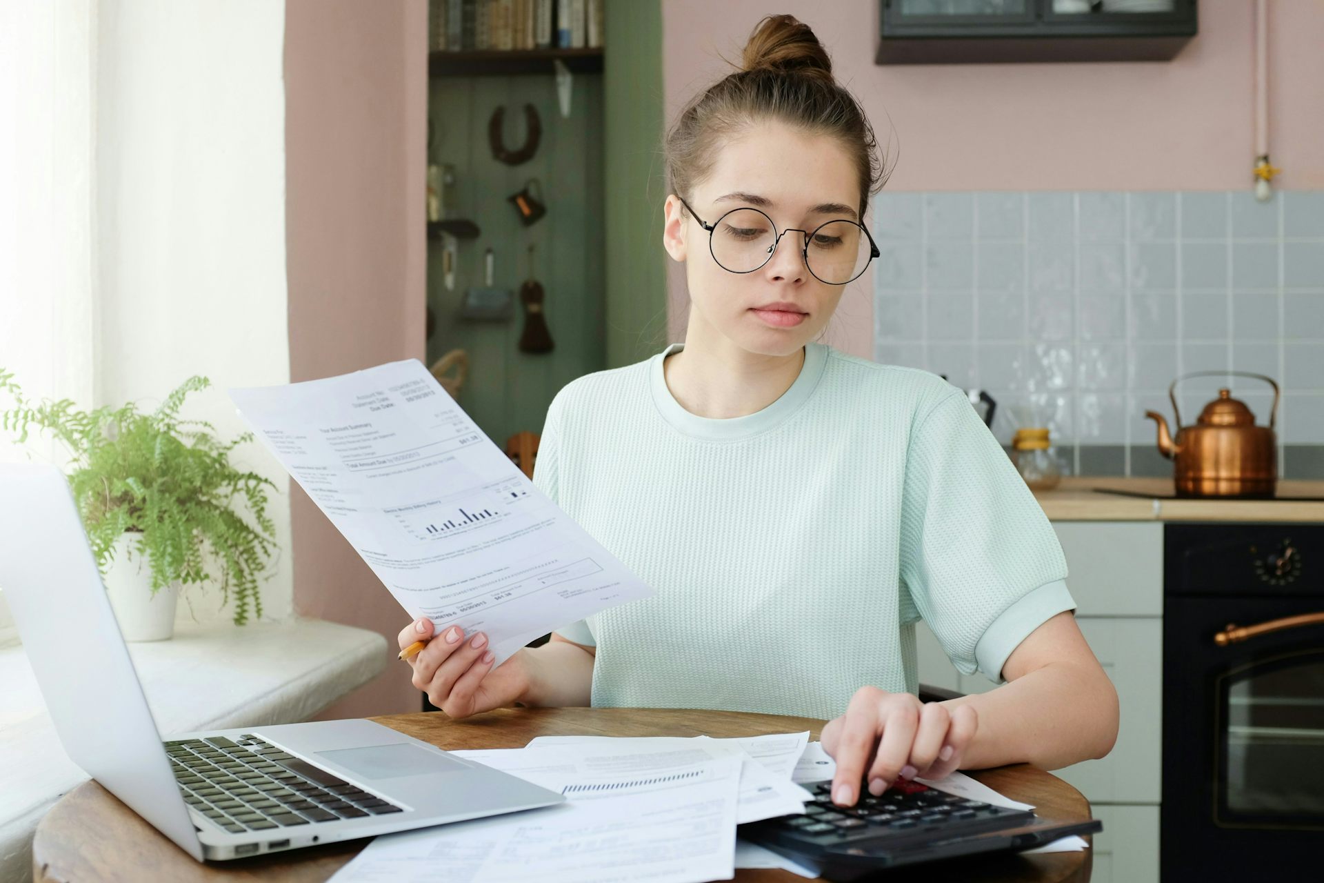 A young woman uses a calculator while sitting at a table in front of a laptop