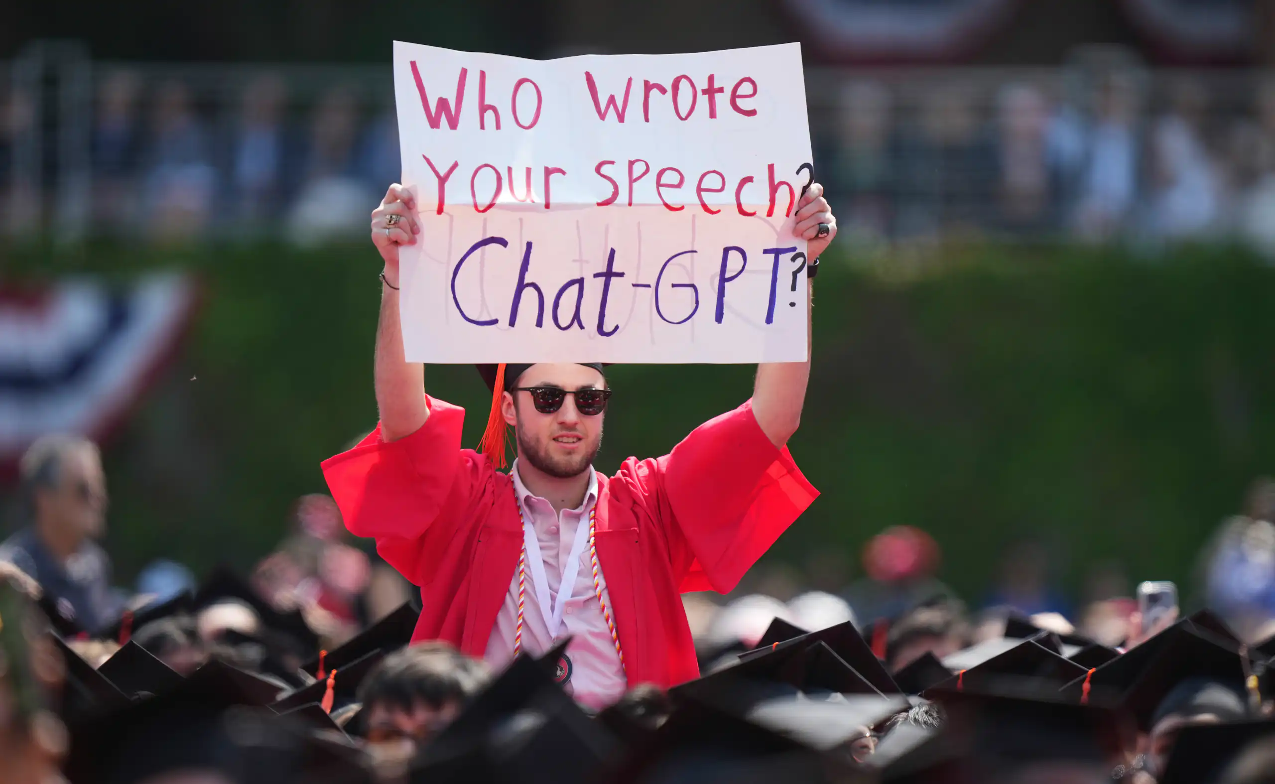 A student at a protest in a red graduation robe and cap holds up a poster with the words who wrote your speech, chat gpt?