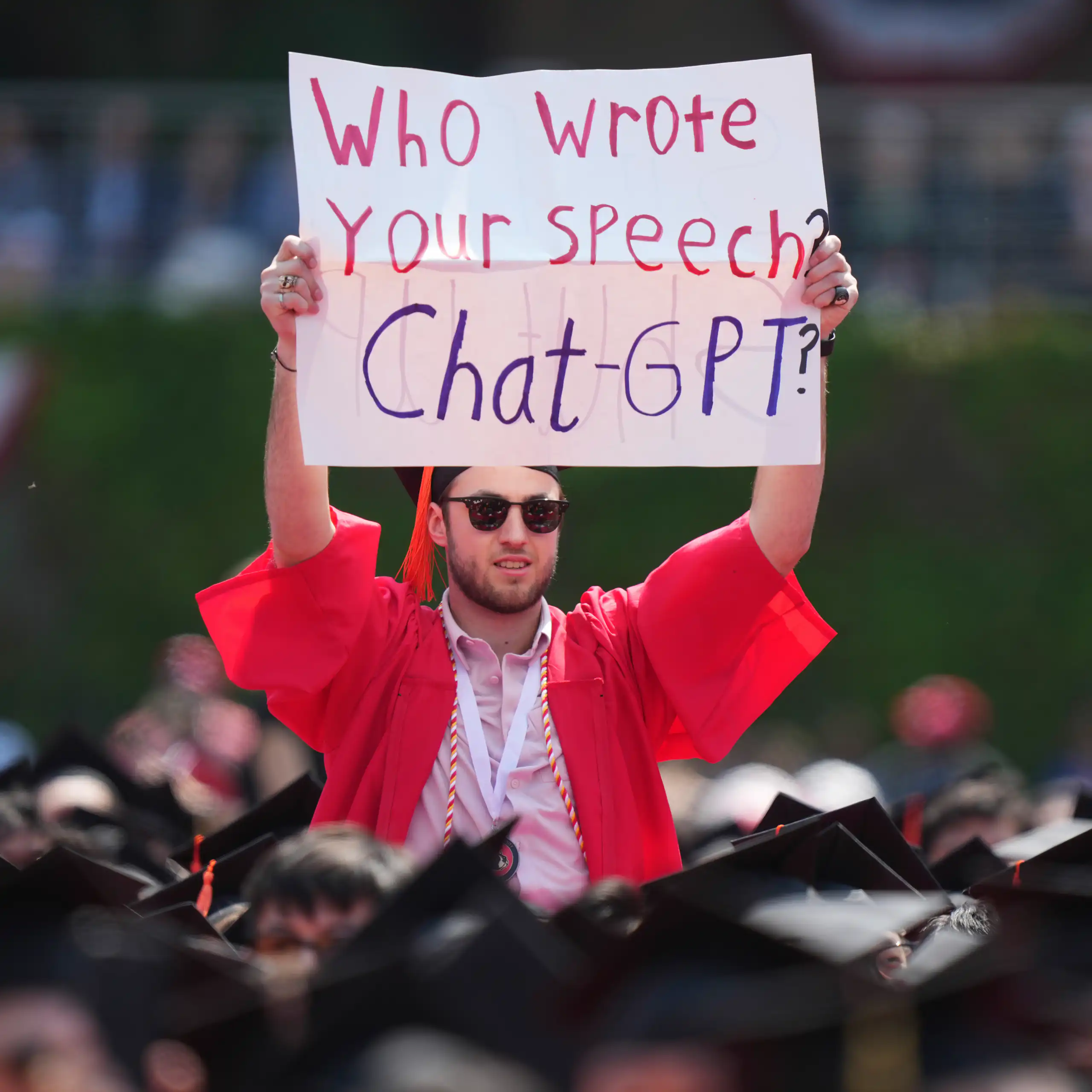 A student at a protest in a red graduation robe and cap holds up a poster with the words who wrote your speech, chat gpt?