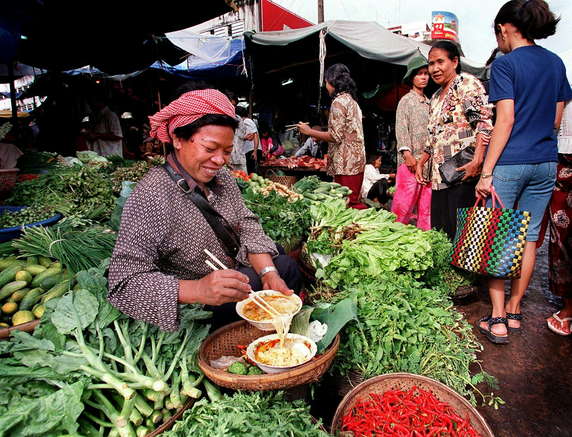 Para pembeli berlalu di dekat seorang pedagang Kamboja yang duduk di tengah hamparan beragam sayur-mayur.