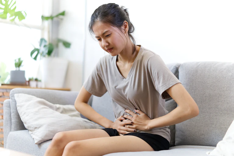 Une jeune fille assise sur un canapé se tient le ventre avec ses deux mains.