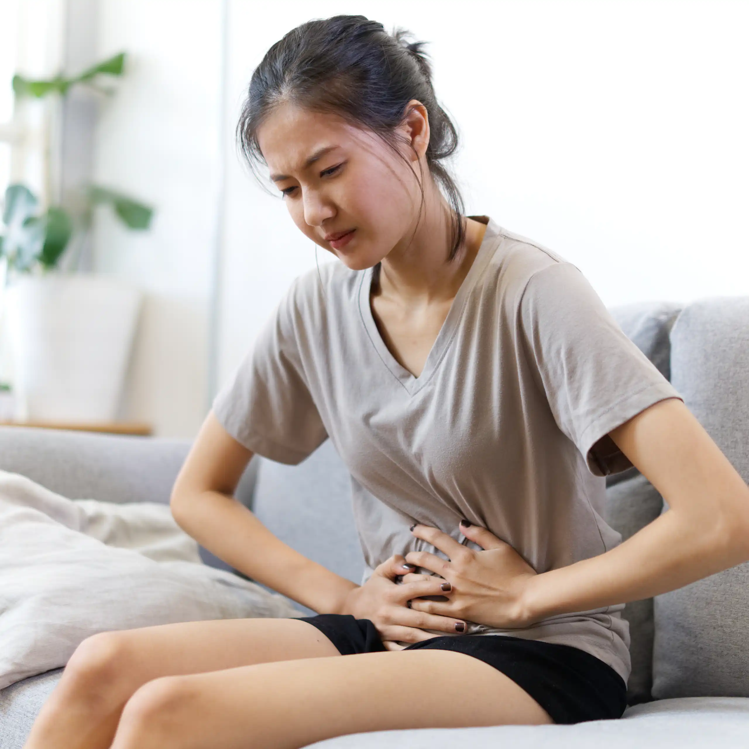 Une jeune fille assise sur un canapé se tient le ventre avec ses deux mains.