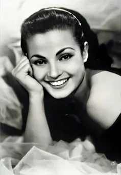 Fotografía en blanco y negro de una mujer joven, sonriente, que apoya la mano en la cara mientras mira a cámara.