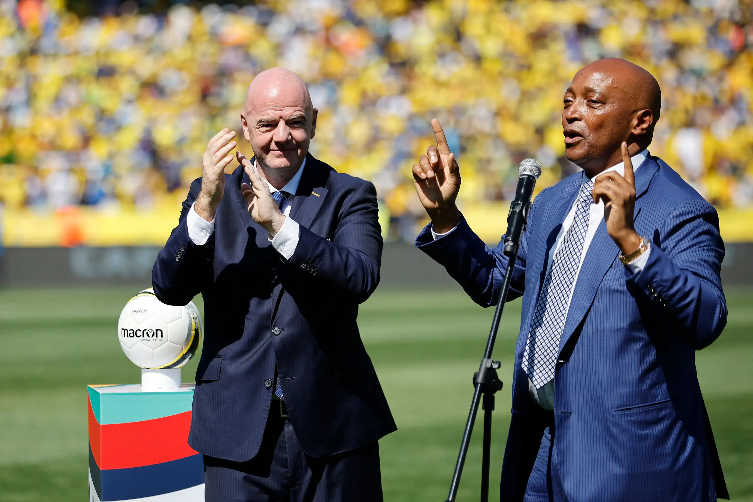 An older, bald-headed European man in a black suit applauds as an African man in a blue suit speaks into a microphone and gestures with his hands, pointing upwards. They're on a grass pitch with crowds in the background.