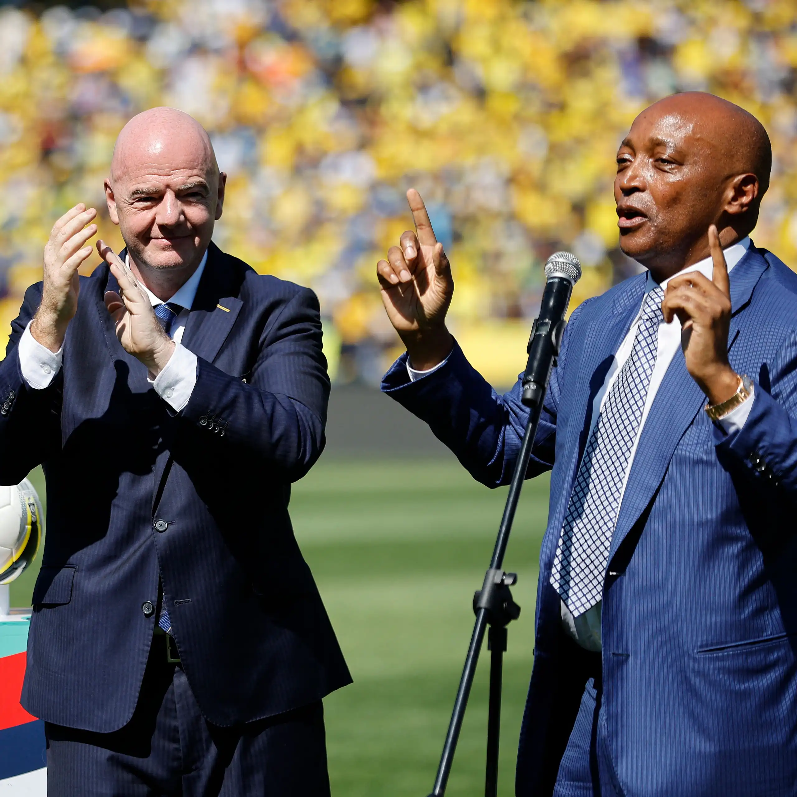 An older, bald-headed European man in a black suit applauds as an African man in a blue suit speaks into a microphone and gestures with his hands, pointing upwards. They're on a grass pitch with crowds in the background.
