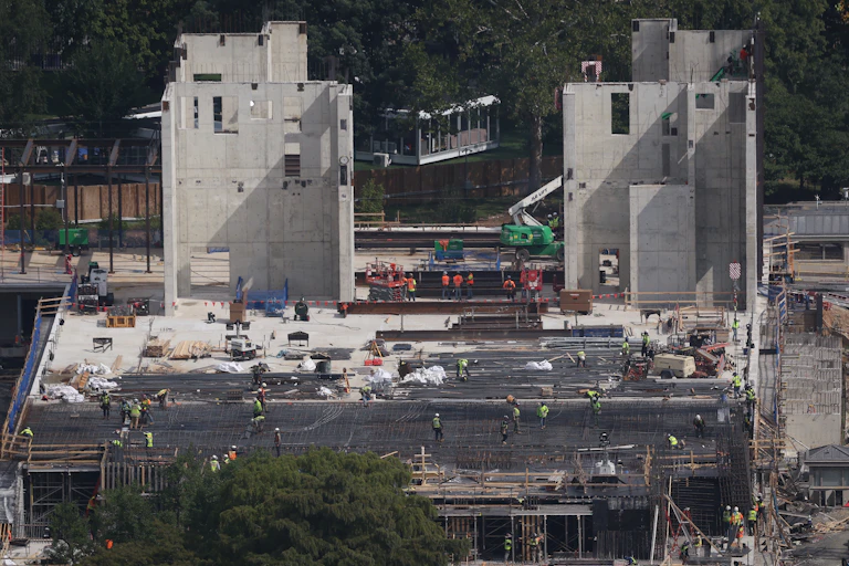 Two large protrusions made of concrete stand atop a large construction project with many workers milling about.