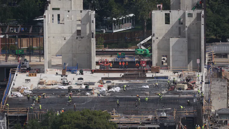 Two large protrusions made of concrete stand atop a large construction project with many workers milling about.