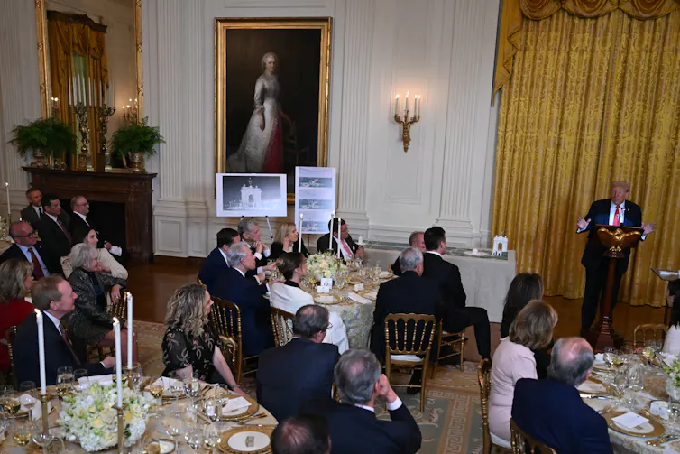 Donald Trump speaks to people in a ballroom with gold-framed paintings.