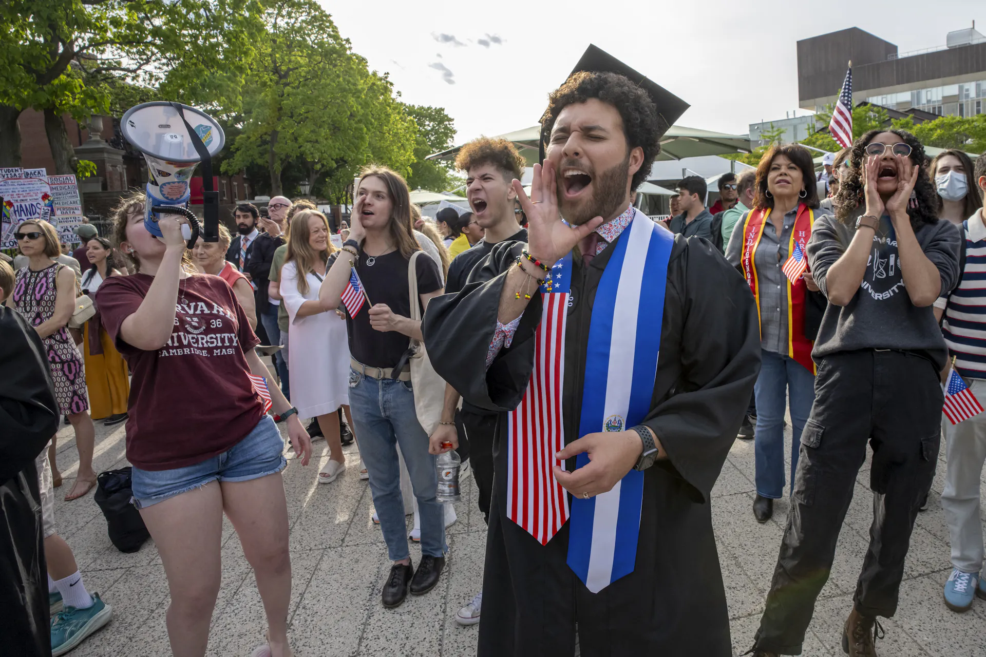 A man with a black robe and graduation cap stands near a group of people shouting with their hands covering their mouths.