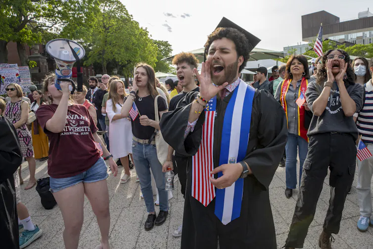 A man with a black robe and graduation cap stands near a group of people shouting with their hands covering their mouths.