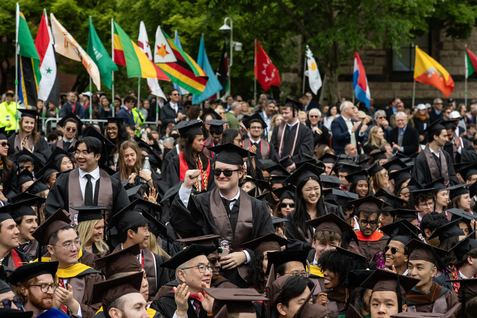 A man with sunglasses and a black graduation cap, gown and sunglasses stands and smiles amid a large crowd of people also wearing black graduation robes. Various colorful country flags wave nearby them.