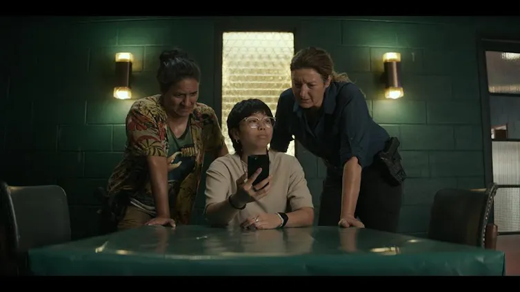 Two woman look over the shoulders of a younger person, seated, holding a smartphone. The one on the right is in a police uniform