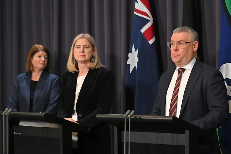 Three politicians wearing suits stand at podiums in front of the Australian flag.