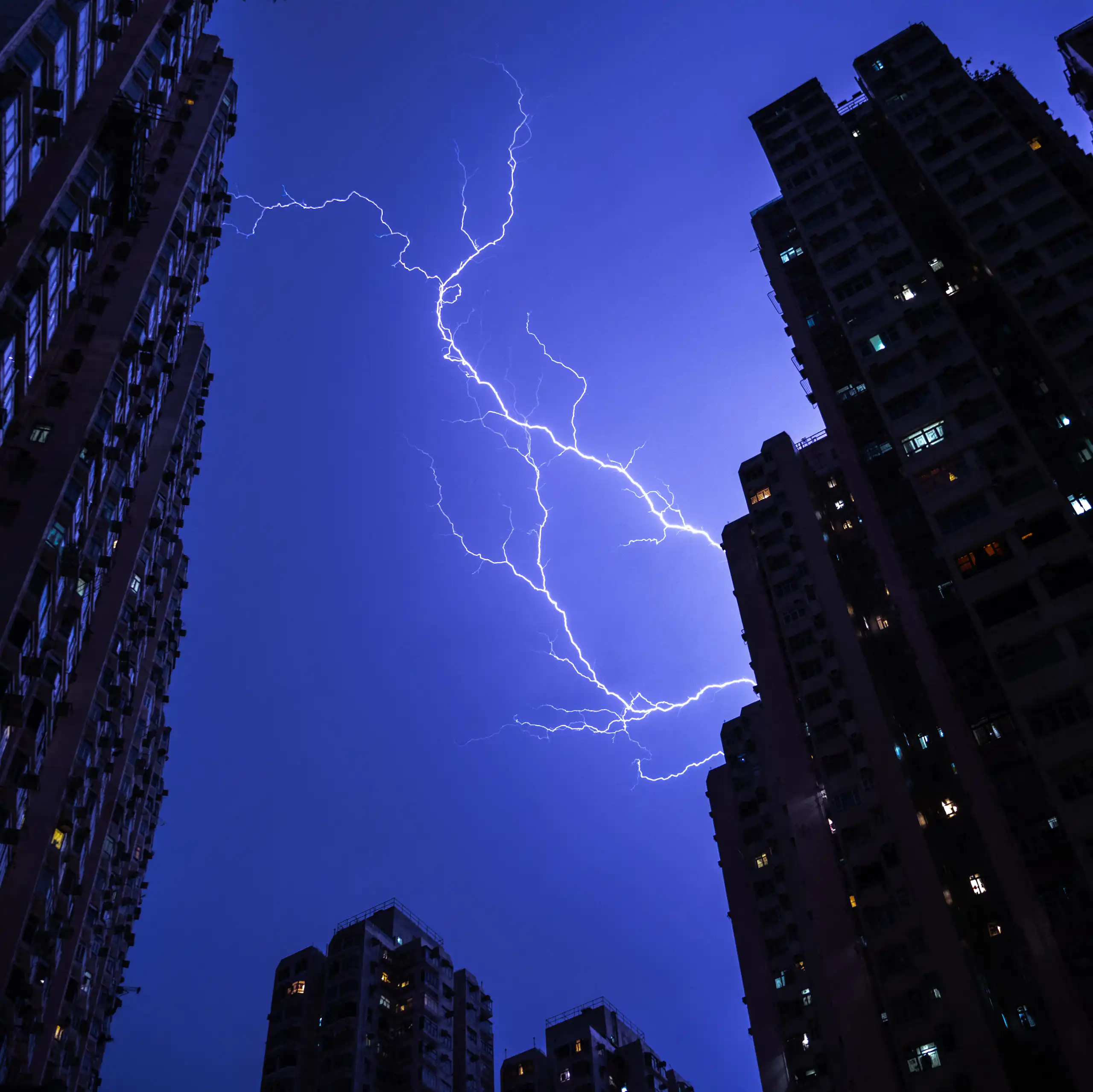Lightning turns the sky purple over a city, with buildings in the foreground and apartment lights on.