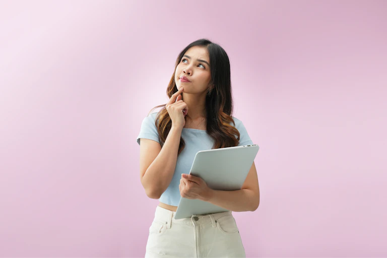 woman standing with laptop, looks questioning, pink background
