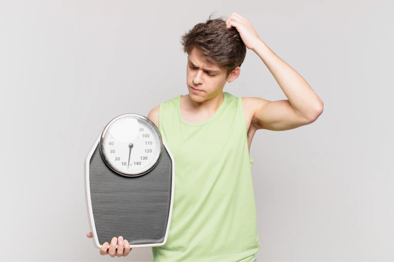 A young man, who is holding a scale in one hand, scratches his head while looking at it in confusion.